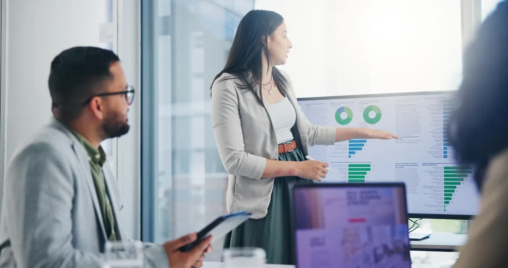 Woman presenting charts on a screen in a meeting room with two colleagues listening attentively
