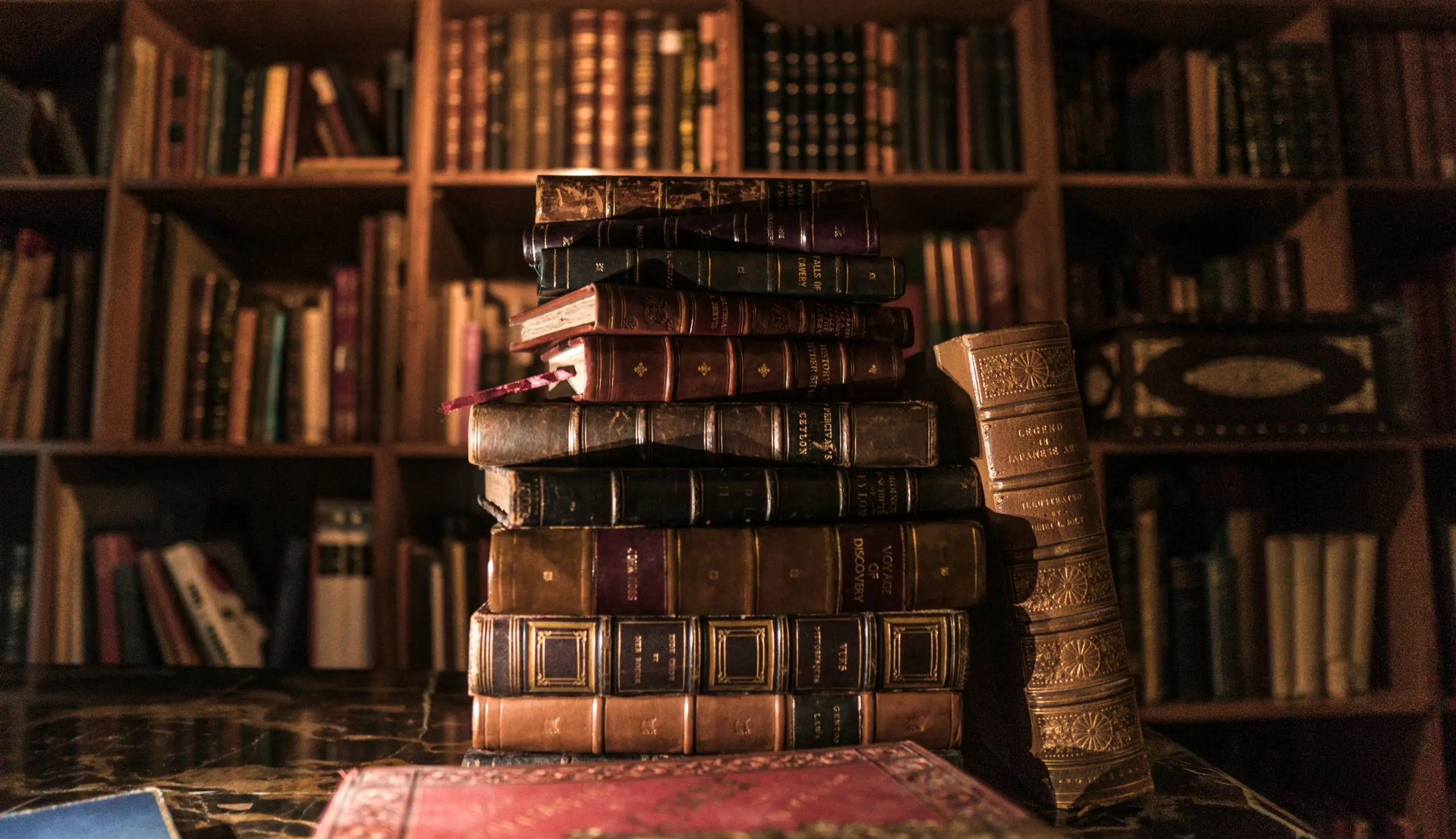 Stack of vintage books on a table with a backdrop of bookshelves filled with more books in a warmly lit library